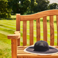 Ein schwarzer Sonnenhut mit breiter Krempe liegt auf einer Gartenbank „Teak Wood Turnberry Bench“ von Alexander Rose (129 cm) in einem sonnigen Garten mit gepflegtem Rasen und Bäumen im Hintergrund.