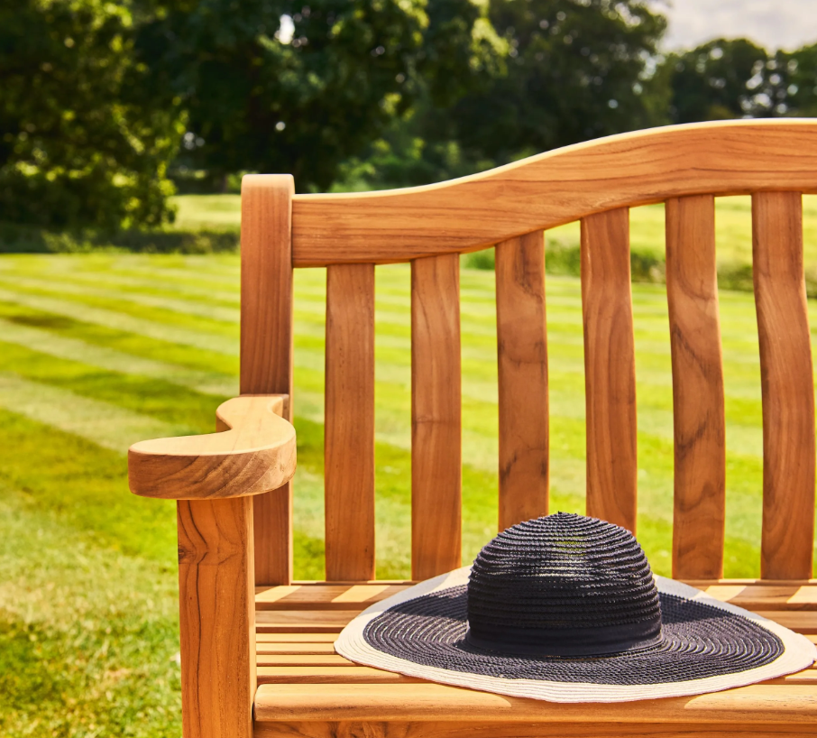 Ein schwarzer Sonnenhut mit breiter Krempe liegt auf einer Gartenbank „Teak Wood Turnberry Bench“ von Alexander Rose (129 cm) in einem sonnigen Garten mit gepflegtem Rasen und Bäumen im Hintergrund.