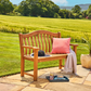 Eine Gartenbank „Teak Wood Turnberry Bench“ von Alexander Rose (129 cm) mit einem rosa Kissen und einer weißen Decke steht auf einer Steinterrasse mit Blick ins Grüne. Ein Hut, ein Glas Wasser und ein Snack in der Nähe schaffen eine einladende Alexander-Rose-Outdoor-Atmosphäre.