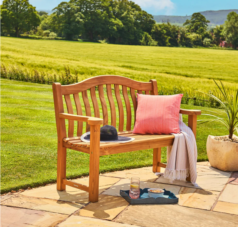 Eine Gartenbank „Teak Wood Turnberry Bench“ von Alexander Rose (129 cm) mit einem rosa Kissen und einer weißen Decke steht auf einer Steinterrasse mit Blick ins Grüne. Ein Hut, ein Glas Wasser und ein Snack in der Nähe schaffen eine einladende Alexander-Rose-Outdoor-Atmosphäre.