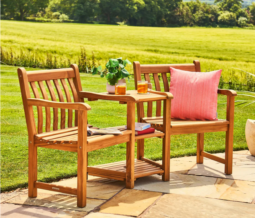 Das „Broadfield Companion Set“ von Alexander Rose aus Teakholz umfasst zwei Gartenstühle aus Premium-Teakholz und einen dazugehörigen Tisch, ausgestattet mit einem rosa Kissen, Platz für Bücher unten und Ablageflächen für Getränke und eine Topfpflanze oben – alles von Alexander Rose.
