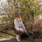 Eine Frau, die den „Esschert Gartenhut Bucket Hat Unisex | Leichter Sonnenhut“ trägt, dazu eine gestreifte Bluse und eine dunkle Hose, hockt auf einem sonnenbeschienenen Gartenweg, umgeben von kahlen, gewunden verzweigten Sträuchern und trockenem Gras.