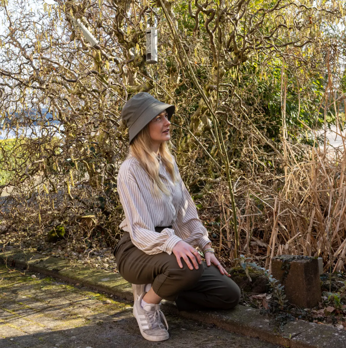 Eine Frau, die den „Esschert Gartenhut Bucket Hat Unisex | Leichter Sonnenhut“ trägt, dazu eine gestreifte Bluse und eine dunkle Hose, hockt auf einem sonnenbeschienenen Gartenweg, umgeben von kahlen, gewunden verzweigten Sträuchern und trockenem Gras.
