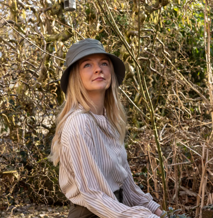 Eine Frau mit langen blonden Haaren, die ein gestreiftes Shirt und einen „Esschert Gartenhut Bucket Hat Unisex | Leichter Sonnenhut“ trägt, sitzt im Freien inmitten kahlgeblätterter Äste und blickt nachdenklich nach oben, während das Sonnenlicht durch die Bäume fällt und einen warmen Schein wirft.