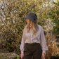 Eine Person mit langen blonden Haaren steht draußen in einem sonnendurchfluteten Garten und trägt den „Esschert Gartenhut Bucket Hat Unisex | Leichter Sonnenhut“ zusammen mit einem gestreiften Hemd und einer braunen Hose – was den entspannten Stil dieses klassischen Hutes perfekt zur Geltung bringt.