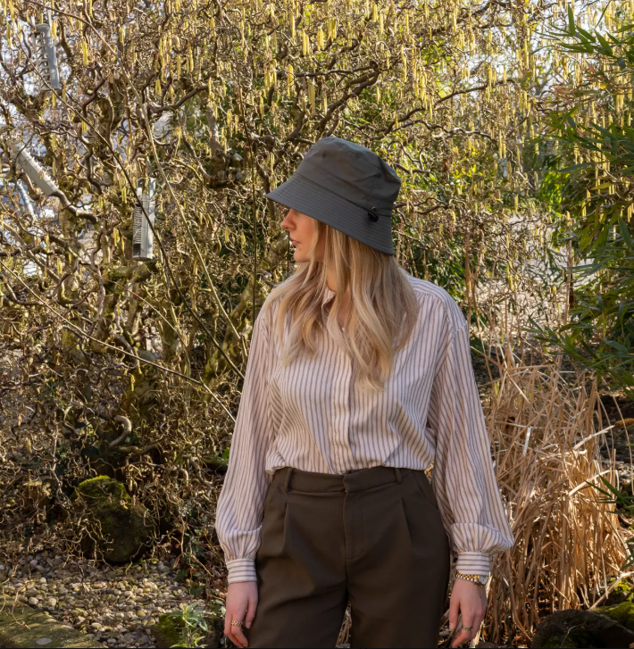 Eine Person mit langen blonden Haaren steht draußen in einem sonnendurchfluteten Garten und trägt den „Esschert Gartenhut Bucket Hat Unisex | Leichter Sonnenhut“ zusammen mit einem gestreiften Hemd und einer braunen Hose – was den entspannten Stil dieses klassischen Hutes perfekt zur Geltung bringt.