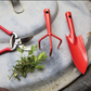Auf einer Metalloberfläche liegen eine rote Gartenschere, ein kleiner roter Handrechen und die „Esschert Kleine Blumenkelle aus Stahl | Rote Pflanzkelle 20,2 cm“ neben einem Zweig mit grünen Blättern – perfekte Gartenwerkzeuge für die Pflege Ihrer Zimmerpflanzen.