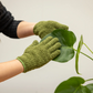 Eine Person, die Esschert-Pflanzenhandschuhe aus Mikrofaser zur Blattpflege in Grün trägt, wischt vorsichtig das große Blatt einer Zimmerpflanze – möglicherweise einer Monstera – mit behutsamen Reinigungstechniken vor einem schlichten, hellen Hintergrund ab.
