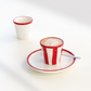 Ein Cappuccino, serviert in der Tasse „Zafferano | Lido Righe Tasse Rot 7 cl“ von Zafferano, die sich durch ihr markantes rot-weiß gestreiftes Design auszeichnet, dazu die passende Untertasse mit Löffel; im Hintergrund ist eine leere weiße Tasse mit rotem Rand zu sehen – alles auf einer weißen Unterlage angeordnet.