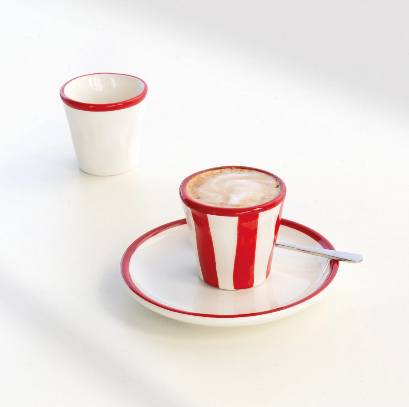 Ein Cappuccino, serviert in der Tasse „Zafferano | Lido Righe Tasse Rot 7 cl“ von Zafferano, die sich durch ihr markantes rot-weiß gestreiftes Design auszeichnet, dazu die passende Untertasse mit Löffel; im Hintergrund ist eine leere weiße Tasse mit rotem Rand zu sehen – alles auf einer weißen Unterlage angeordnet.