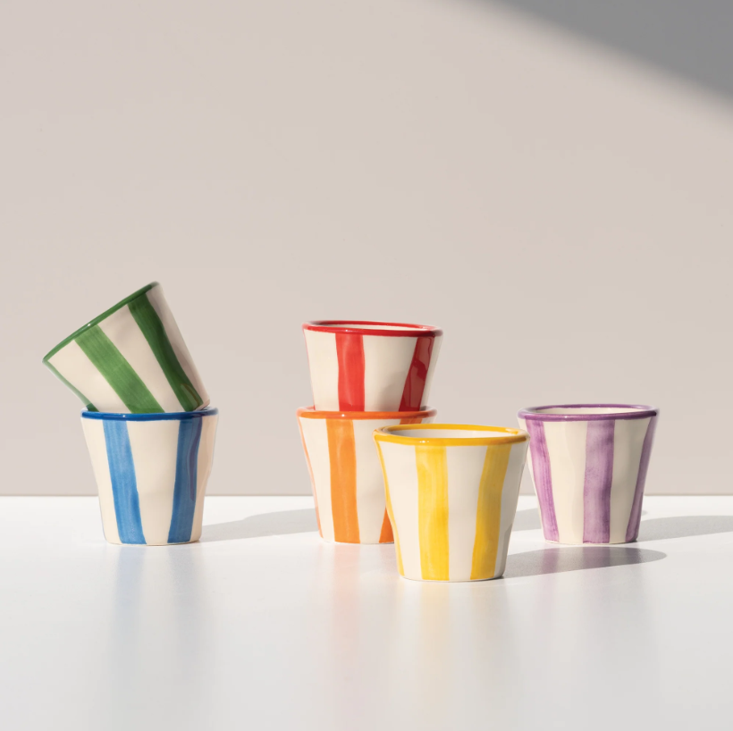 Sechs Keramikbecher der Serie „Zafferano Lido Righe Tasse Gelb“, die jeweils mit kräftigen vertikalen Streifen in leuchtenden Grün-, Rot-, Orange-, Gelb- und Violetttönen verziert sind, stehen auf einer weißen Fläche angeordnet, wobei zwei davon übereinander gestapelt sind; sie verkörpern den lebendigen Geist des italienischen Designs, der auch die klassischen Zafferano-Espressotassen auszeichnet, und fangen das Sonnenlicht ein, das im Hintergrund sanfte Schatten wirft.