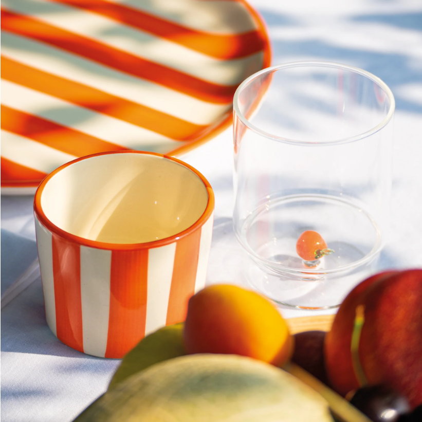 Eine lebendige Szene mit der „Zafferano | Lido Righe Tasse oder Schale Orange 23 cl“ von Zafferano, elegant kombiniert mit einem orange-weiß gestreiften Teller, einem klaren Glas mit einer einzelnen Kirschtomate und verschiedenen Früchten auf einer weißen Tischdecke, die von fleckigem Sonnenlicht beleuchtet wird. Eine italienische Keramikschale rundet das fröhliche Ambiente mit handwerklichem Flair ab.