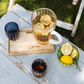Auf einem Tablett steht ein Krug Eistee mit Zitronenscheiben, dazu zwei blaue „Zafferano | Perle Glas Blau“-Becher (32 cl), die in der Tradition der italienischen Glasmacherkunst meisterhaft gefertigt wurden. Daneben steht eine grüne Schale mit Zitronen und Zitronenscheiben auf einem rustikalen Tisch im Freien, im Hintergrund ist Gras zu sehen.