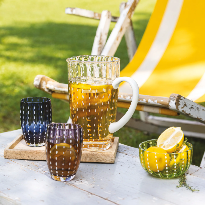Auf einem rustikalen Tisch steht ein Glaskrug mit Eistee, daneben zwei Gläser „Zafferano Perle Glas Blau 32 cl“ von Zafferano, gefertigt in echter italienischer Glasmacherkunst und mundgeblasen, sowie eine Schale mit Zitronen – neben einem gelb gestreiften Liegestuhl auf grünem Gras im Freien.