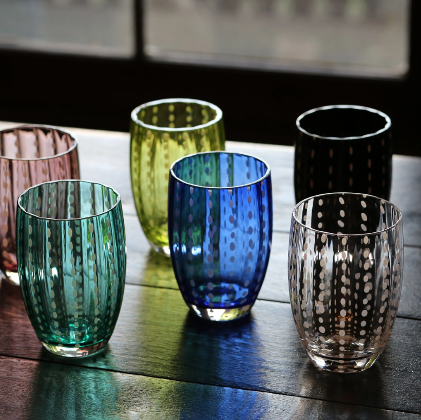 Sechs farbenfrohe Zafferano Perle-Gläser, darunter das Zafferano | Perle Glas Blau 32 cl, stehen in zwei ordentlichen Reihen auf einem Holztisch. Jedes Glas hat eine andere Farbe – Rosa, Gelb, Grün, Blau, Schwarz und klar – und ist mit verspielten gepunkteten Streifen verziert, die die exquisite italienische Handwerkskunst unterstreichen. Diese unverwechselbaren Zafferano-Stücke aus mundgeblasenem Glas verleihen jeder Umgebung künstlerisches Flair und Farbe.