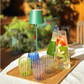 Ein Holztablett mit vier farbenfrohen, gepunkteten Gläsern – Zafferano | Perle Glas Blau 32 cl von Zafferano, gefertigt in exquisiter italienischer Glasmacherkunst aus mundgeblasenem Glas –, eine kleine grüne Lampe, eine Flasche mit grüner Flüssigkeit und eine Karaffe mit Wasser, Erdbeeren und Minze, alles auf einem sonnendurchfluteten Tisch im Freien angeordnet.