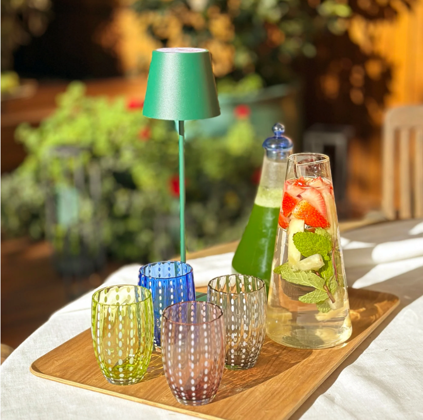 Ein Holztablett mit vier farbenfrohen, gepunkteten Gläsern – Zafferano | Perle Glas Blau 32 cl von Zafferano, gefertigt in exquisiter italienischer Glasmacherkunst aus mundgeblasenem Glas –, eine kleine grüne Lampe, eine Flasche mit grüner Flüssigkeit und eine Karaffe mit Wasser, Erdbeeren und Minze, alles auf einem sonnendurchfluteten Tisch im Freien angeordnet.