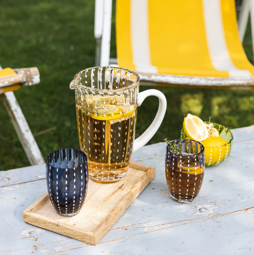 Auf einem Tisch im Freien steht ein Glaskrug mit Eistee und Zitronenscheiben auf einem Holztablett, daneben zwei Gläser der Serie „Zafferano | Perle Glas Blau“ (32 cl) – beeindruckende Beispiele italienischer Glasmacherkunst. Im Hintergrund sind eine Schale mit Zitronen und ein gelb gestreifter Stuhl zu sehen.