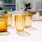 Ein Set aus vier „Zafferano | Perle Glas Amber“-Gläsern (32 cl) – meisterhaft von Zafferano in der Tradition der feinen italienischen Glasmacherkunst handgefertigt – mit jeweils einem einzigartigen Strukturmuster, angeordnet auf einer weißen Fläche im Außenbereich. Diese aus mundgeblasenem Glas gefertigten, bernsteinfarbenen Stücke werfen im Sonnenlicht eindrucksvolle Schatten und heben sich lebhaft von einem sanft unscharfen Hintergrund ab.