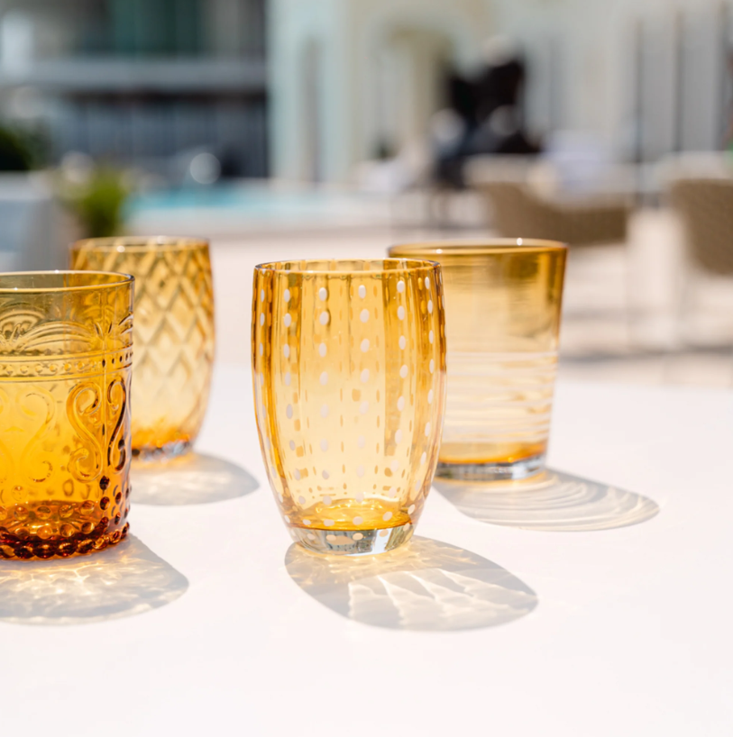 Ein Set aus vier „Zafferano | Perle Glas Amber“-Gläsern (32 cl) – meisterhaft von Zafferano in der Tradition der feinen italienischen Glasmacherkunst handgefertigt – mit jeweils einem einzigartigen Strukturmuster, angeordnet auf einer weißen Fläche im Außenbereich. Diese aus mundgeblasenem Glas gefertigten, bernsteinfarbenen Stücke werfen im Sonnenlicht eindrucksvolle Schatten und heben sich lebhaft von einem sanft unscharfen Hintergrund ab.