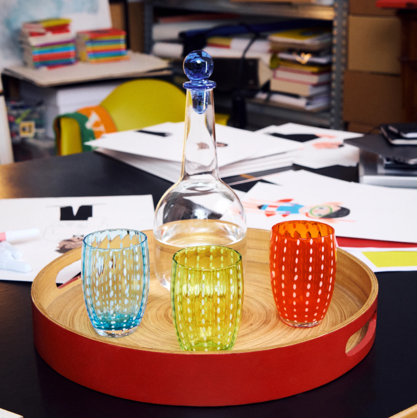 Auf einem Tisch, der von Papieren, Büchern und Büromaterial umgeben ist, steht ein Holztablett mit einer Glaskaraffe und drei farbenfrohen Gläsern – darunter das „Zafferano | Perle Glas Apfelgrün 32 cl“. Jedes Glas von Zafferano zeugt durch seine mundgeblasene Kunstfertigkeit von italienischer Handwerkskunst und elegantem Design.