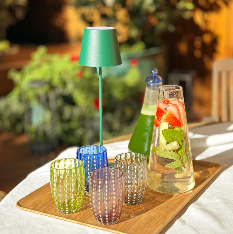 Auf einem Holztablett stehen eine grüne Lampe, vier leuchtend grüne „Zafferano | Perle Glas Apfelgrün“-Becher (32 cl) – eindrucksvolle Beispiele italienischer Glasherstellung von Zafferano – sowie eine Karaffe, gefüllt mit Wasser, Erdbeeren, Minze und Limette. Außerdem ist eine zweite Flasche mit einer grünen Flüssigkeit zu sehen. Sonnenlicht strömt über die Szene, im Hintergrund sind üppige Pflanzen zu sehen.