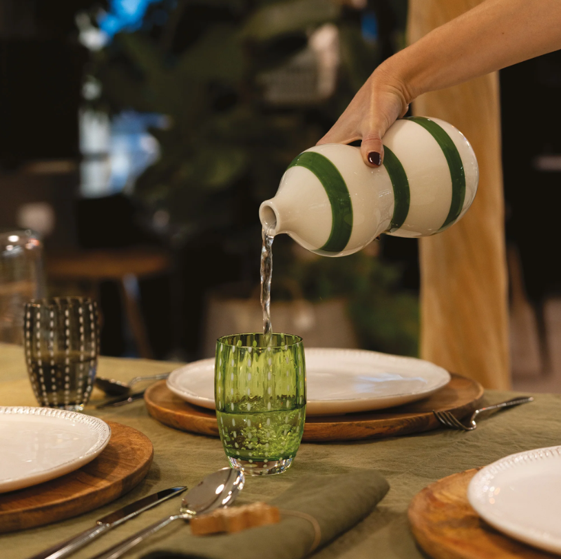 Eine Hand gießt Wasser aus einem gestreiften Krug in ein „Zafferano | Perle Glas Apfelgrün 32 cl“ auf einem gedeckten Esstisch mit Tellern, Besteck und Holzuntertellern, das die exquisite italienische Glasmacherkunst zur Geltung bringt.