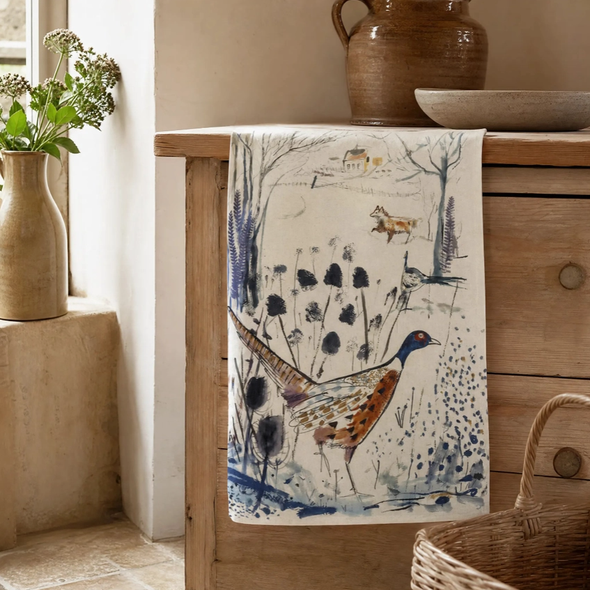 Ein Sam Wilson-Küchentuch „Pheasant & Fox“ aus 100 % Baumwolle, in England hergestellt und mit einem handgemalten Fasan- und Fuchs-Motiv verziert, hängt an einem Holzschrank. In unmittelbarer Nähe stehen rustikale Keramik, eine dekorative Vase mit frischem Grün und ein geflochtener Korb in einem gemütlichen, sonnendurchfluteten Raum.