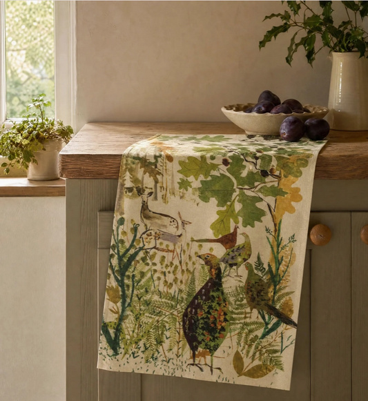 Ein Sam Wilson-Küchentuch „Pheasant & Deer“ aus 100 % Baumwolle, hergestellt in England, hängt über der Küchenzeile neben einem Fenster und verleiht dem Raum den Charme des britischen Landhausstils. Auf der Holzarbeitsplatte stehen eine Schale mit dunklen Pflaumen und eine Vase mit grünen Zweigen.
