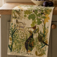 Ein Sam-Wilson-Küchentuch „Pheasant & Deer“ aus 100 % in England hergestellter Baumwolle mit handgezeichnetem Fasanen-, Hirsch- und Blattmotiv hängt über einem Holzschrank – ein stilvolles Accessoire im britischen Landhausstil. Auf der Arbeitsplatte darüber steht eine Schale mit dunklen Pflaumen.