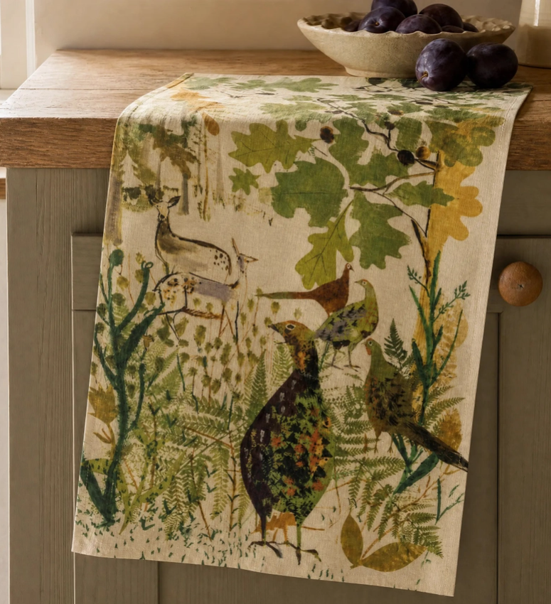 Ein Sam-Wilson-Küchentuch „Pheasant & Deer“ aus 100 % in England hergestellter Baumwolle mit handgezeichnetem Fasanen-, Hirsch- und Blattmotiv hängt über einem Holzschrank – ein stilvolles Accessoire im britischen Landhausstil. Auf der Arbeitsplatte darüber steht eine Schale mit dunklen Pflaumen.