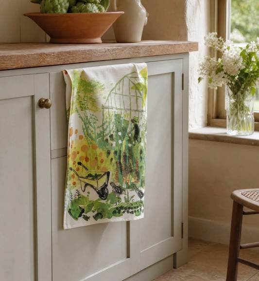 An einem hellgrauen Schrank hängt ein Sam-Wilson-Küchentuch „Greenhouse“ aus 100 % in England hergestellter Baumwolle mit einem charmanten Gewächshausmotiv. Auf der Arbeitsfläche am sonnendurchfluteten Fenster stehen eine Holzschale mit grünen Äpfeln und eine Vase mit weißen Blumen.