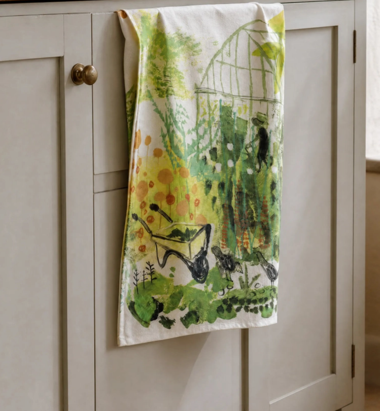 Ein Küchentuch mit einer grünen Gartenillustration – komplett mit einer Schubkarre und einem bezaubernden Gewächshausmotiv – hängt an einer hellen Schranktür mit runden Messingknöpfen. Das Sam Wilson Küchentuch „Greenhouse“, hergestellt in England aus 100 % Baumwolle, bringt Stil und Funktionalität in Ihre Küche.