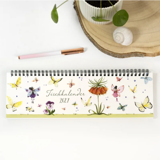 Ein spiralgebundener Tischkalender „Schmetterlinge 2027“ des Grätz Verlags mit verspielten Schmetterlingsillustrationen von Silke Leffler liegt auf einer weißen Unterlage neben einem rosa Stift, einer Topfpflanze, einer Holzscheibe und einer weißen Muschel – und ist damit sowohl ein bezaubernder Wochenplaner als auch ein dekoratives Highlight für jeden Arbeitsplatz.