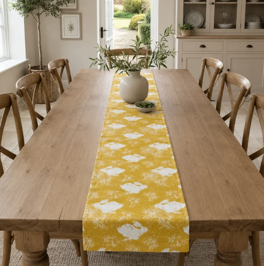 Ein Esstisch aus Holz ist mit dem Tischläufer „Sam Wilson | Headlong Hare“ in der Farbe „Yellow Ochre“ (34 × 196 cm) geschmückt, der gelbe und weiße Muster aufweist. Auf dem Tischläufer stehen eine weiße Vase mit grünen Zweigen und eine kleine Schale, während Holzstühle den Tisch in einem hellen, luftigen Esszimmer umgeben.