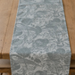 Ein Holztisch mit dem Tischläufer „Headlong Hare“ von Sam Wilson in der Ausführung „Owl & Fox“ (34 × 196 cm), einem graublauen Tischläufer, der mit einem filigranen Waldmotiv aus Bäumen, Blättern und versteckten Tieren wie Eulen und Füchsen verziert ist.