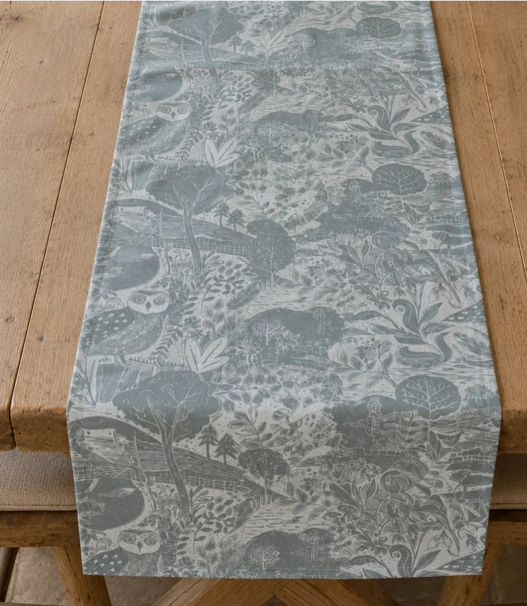 Ein Holztisch mit dem Tischläufer „Headlong Hare“ von Sam Wilson in der Ausführung „Owl & Fox“ (34 × 196 cm), einem graublauen Tischläufer, der mit einem filigranen Waldmotiv aus Bäumen, Blättern und versteckten Tieren wie Eulen und Füchsen verziert ist.