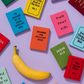 Eine gelbe Banane liegt auf einem lavendelfarbenen Untergrund, umgeben von bunten Miniaturbüchern mit skurrilen Titeln wie „Dream Big“ und „I’d Rather Be at Home With My Dog“ – jedes einzelne spiegelt den eigenwilligen Charakter des „Ohh Deer | ’Can’t Adult Today’ Mini A7 Notizbuch“ von Ohh Deer wider.
