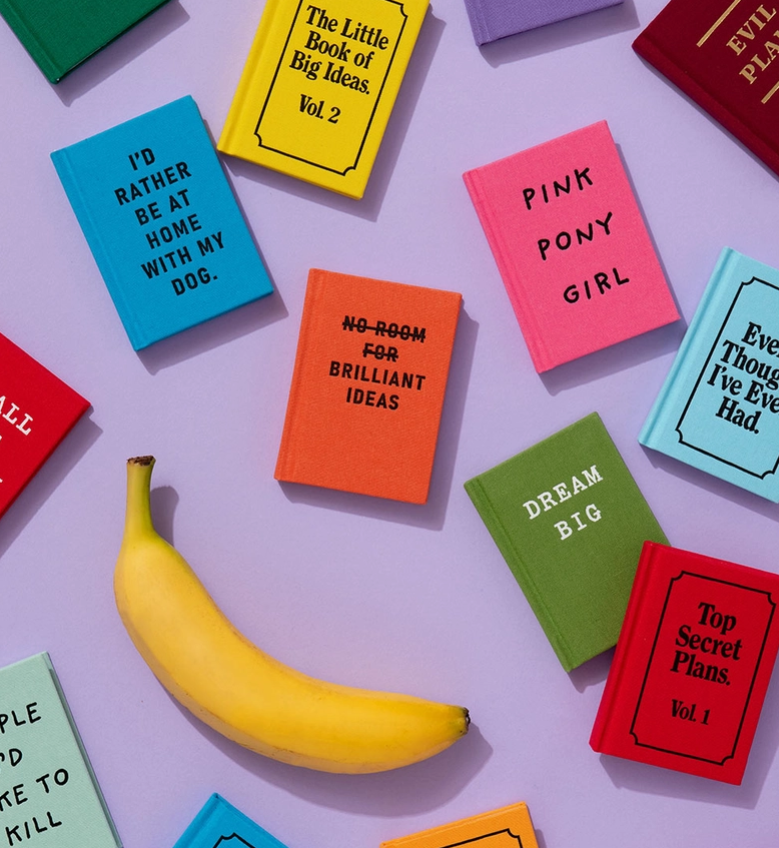 Eine gelbe Banane liegt auf einem lavendelfarbenen Untergrund, umgeben von bunten Miniaturbüchern mit skurrilen Titeln wie „Dream Big“ und „I’d Rather Be at Home With My Dog“ – jedes einzelne spiegelt den eigenwilligen Charakter des „Ohh Deer | ’Can’t Adult Today’ Mini A7 Notizbuch“ von Ohh Deer wider.