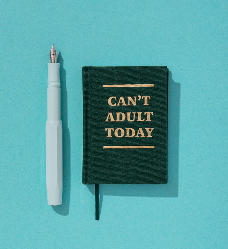 Ein weißer Füllfederhalter und ein Ohh Deer „Can’t Adult Today“-Mini-Notizbuch im Format A7 in Dunkelgrün mit goldener Beschriftung, beide vor blauem Hintergrund.