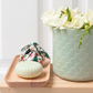 Auf einem Holztablett und einem Tisch vor weißem Hintergrund stehen eine hellgrüne, strukturierte Vase mit weißen Blumen, ein Stück Castelbel-Seife „Chita Verbena“ (150 g) und ein kleiner Stoffbeutel mit Blumenmuster.