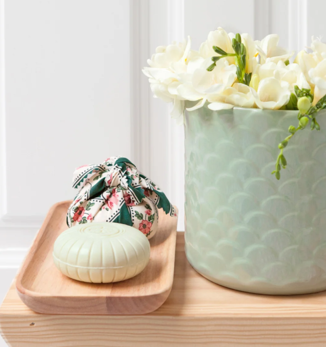 Auf einem Holztablett und einem Tisch vor weißem Hintergrund stehen eine hellgrüne, strukturierte Vase mit weißen Blumen, ein Stück Castelbel-Seife „Chita Verbena“ (150 g) und ein kleiner Stoffbeutel mit Blumenmuster.