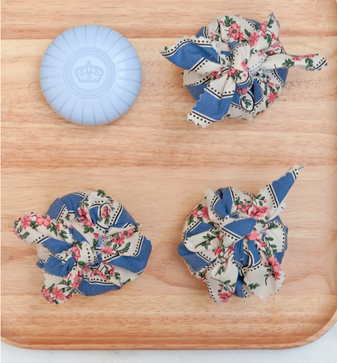 Auf einem Holztablett liegen ein rundes blaues Seifenstück und drei in blaues, mit Blumenmustern bedrucktes Tuch gewickelte Bündel, die den Charme von Castelbel verströmen | Seife Chita Lavender 150 g – eine luxuriöse portugiesische Seife von Castelbel mit eleganten Lavendelnoten.