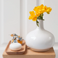 Auf einem Holztisch steht eine weiße Vase mit gelben Blumen neben einem Holztablett, auf dem ein kleiner runder weißer Gegenstand und ein in Stoff gewickeltes Objekt mit Blumenmuster liegen – eine ideale Geschenkidee, besonders in Kombination mit der eleganten Castelbel-Seife „Chita White Jasmine“ (150 g).