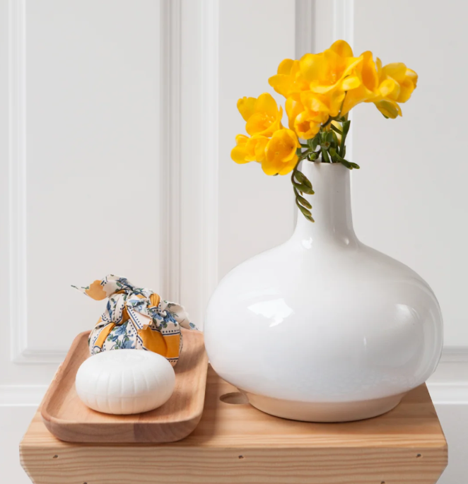Auf einem Holztisch steht eine weiße Vase mit gelben Blumen neben einem Holztablett, auf dem ein kleiner runder weißer Gegenstand und ein in Stoff gewickeltes Objekt mit Blumenmuster liegen – eine ideale Geschenkidee, besonders in Kombination mit der eleganten Castelbel-Seife „Chita White Jasmine“ (150 g).