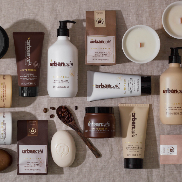 Eine Auswahl an Bade- und Körperpflegeprodukten der Serie „Urban Caffé Mocha“ von Somerset Toiletry – darunter die Handseife „Urban Caffé Mocha“ (500 ml), Seifen, Körperpeelings, Kerzen und Lotionen, die mit Kaffeeextrakt angereichert sind – ist übersichtlich auf einer beigen Leinenunterlage angeordnet und mit Kaffeebohnen akzentuiert.