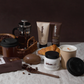 Die Handseife „Urban Caffé Mocha“ (500 ml) von Somerset Toiletry, angereichert mit Kaffeeextrakt, wird kunstvoll neben einer French Press, Kaffeebohnen, Croissants, einer brennenden Kerze und einem Kaffee-To-Go-Becher auf einem Tisch vor einem neutralen Hintergrund präsentiert.