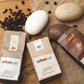 Drei Seifenstücke in Form von Kaffeebohnen – zwei braune und ein weißes – sind zusammen mit der passenden Verpackung „Somerset Toiletry Urban Caffé Mocha Soap Bar 180 g“ und verstreuten Kaffeebohnen auf einer Juteunterlage angeordnet. Diese luxuriösen Handseifen, angereichert mit Kaffeeextrakt und Kakaobutter, fangen das reichhaltige Aroma Ihres Lieblingscafés ein.