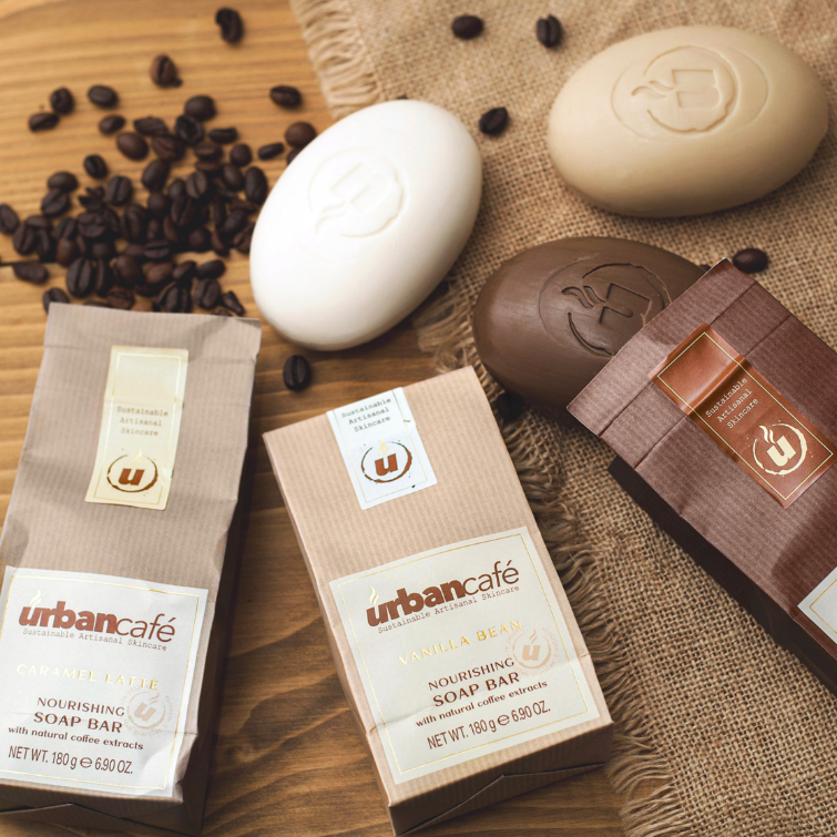 Drei Seifenstücke in Form von Kaffeebohnen – zwei braune und ein weißes – sind zusammen mit der passenden Verpackung „Somerset Toiletry Urban Caffé Mocha Soap Bar 180 g“ und verstreuten Kaffeebohnen auf einer Juteunterlage angeordnet. Diese luxuriösen Handseifen, angereichert mit Kaffeeextrakt und Kakaobutter, fangen das reichhaltige Aroma Ihres Lieblingscafés ein.