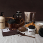 Die Handseife „Urban Caffé Vanilla“ (500 ml) von Somerset Toiletry ist auf einer braunen Fläche neben Kaffeebohnen, Croissants, einer French Press, einer Kerze und Kaffeebechern zum Mitnehmen angeordnet, alles vor einem dunkelbraunen Hintergrund.